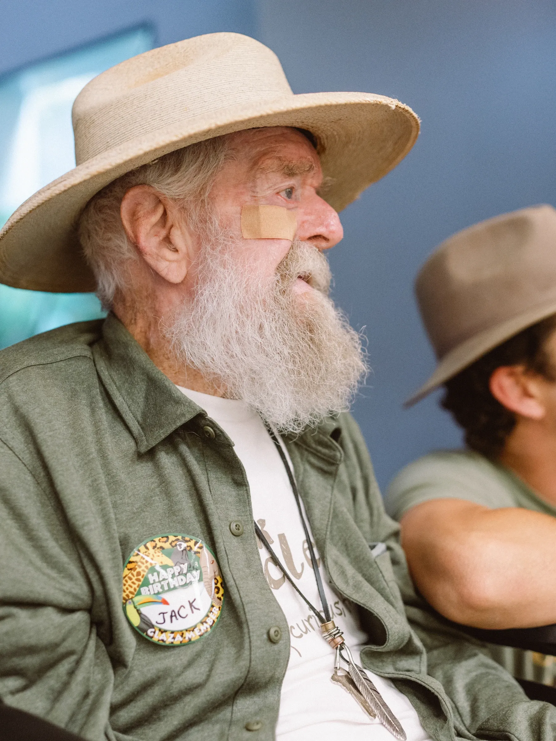 Uncle Jack, in a wide-brimmed hat with a “Happy Birthday Jack” badge on his jacket, sits beside a younger man in a hat.